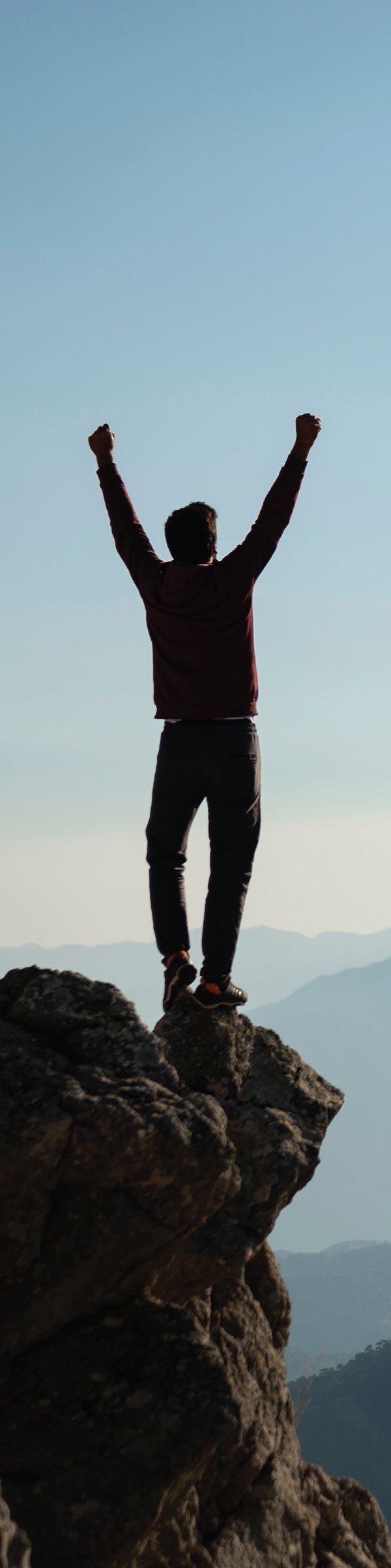 Photograph of a person seen from behind, standing on a rock with both arms raised, mountains and sky beyond. No text.